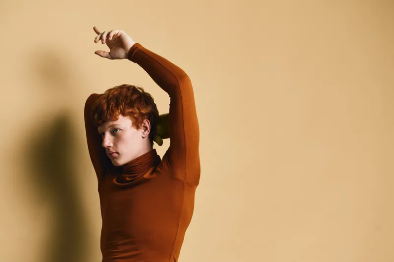Young man with red hair posing in brown turtleneck sweater against beige background with artistic shadow and thoughtful facial expression.
