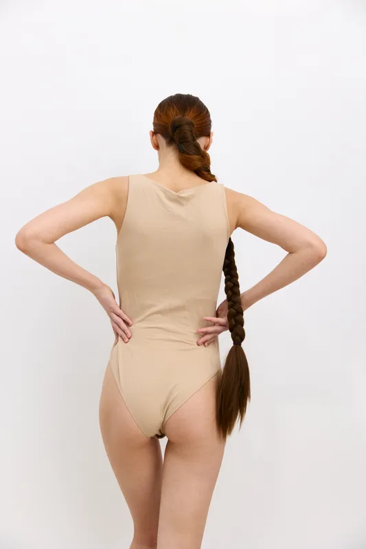 woman with long braided hair wearing beige bodysuit standing with hands on hips against plain white background