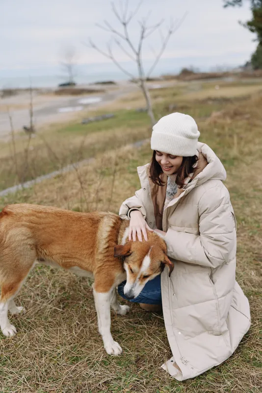 Dog, woman, outdoors, caring, winter, coat, park, nature, companionship, animal in a tranquil park setting with a gentle touch on a calm dog beside a lake under soft light and cloudy sky