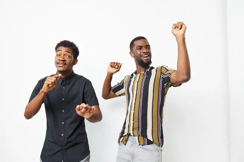 Two joyful African American men dancing with enthusiasm in casual outfits. The bright background highlights their vibrant energy and liveliness, capturing a moment of celebration.