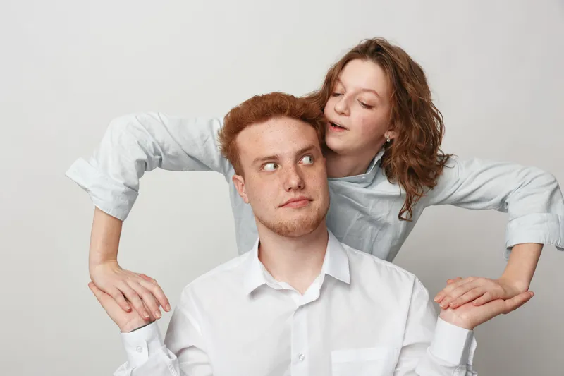 Young redhead man and woman in white shirts hold hands and interact playfully against light background. Concept of friendship, trust, and casual lifestyle with natural expressions.