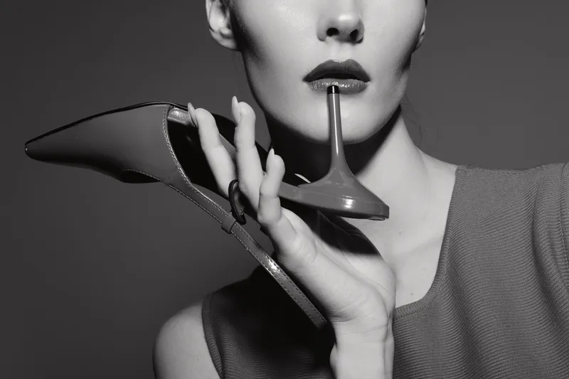 Close-up of white woman with light skin holding high heel shoe near lips in studio. Concept of fashion, elegance, and luxury lifestyle in black and white.