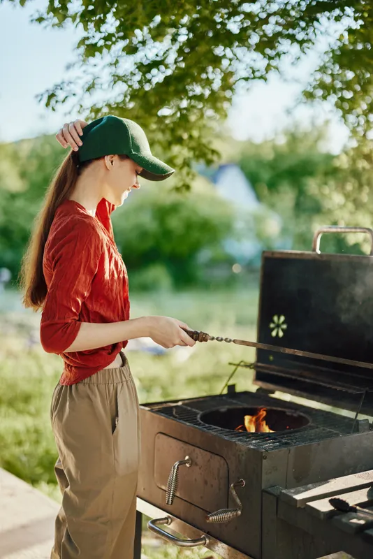 Young woman grilling outdoors, wearing a red shirt and cap, smiling with happiness, surrounded by green nature, representing summer joy and outdoor cooking