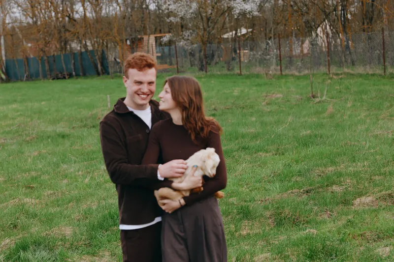 Joyful couple outdoors, smiling while holding a gentle goat kid, warm sunlight, grassy meadow, casual clothing, wholesome moment, natural colors, tranquil countryside backdrop