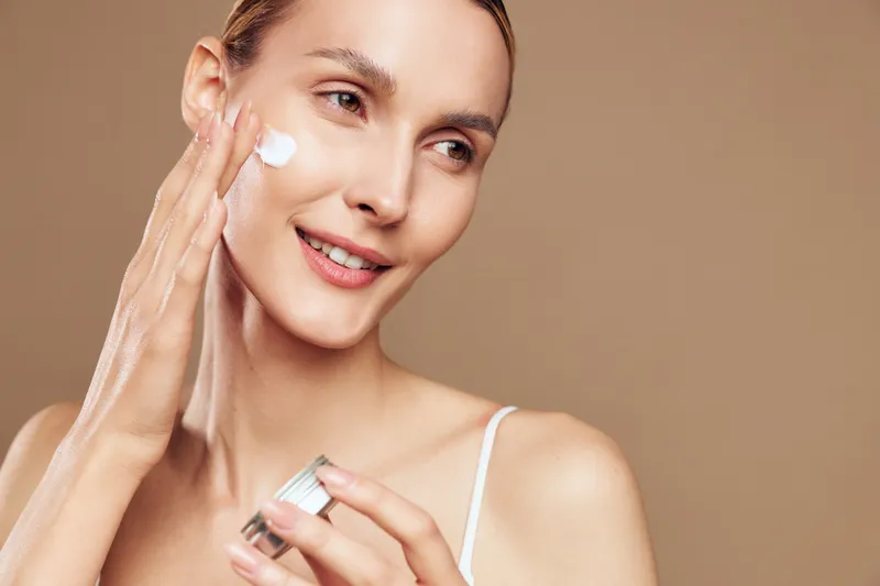 Smiling young woman applying moisturizer cream to face, close up beauty portrait, spa inspired self care routine, smooth hydrated skin and natural glow, beige studio background