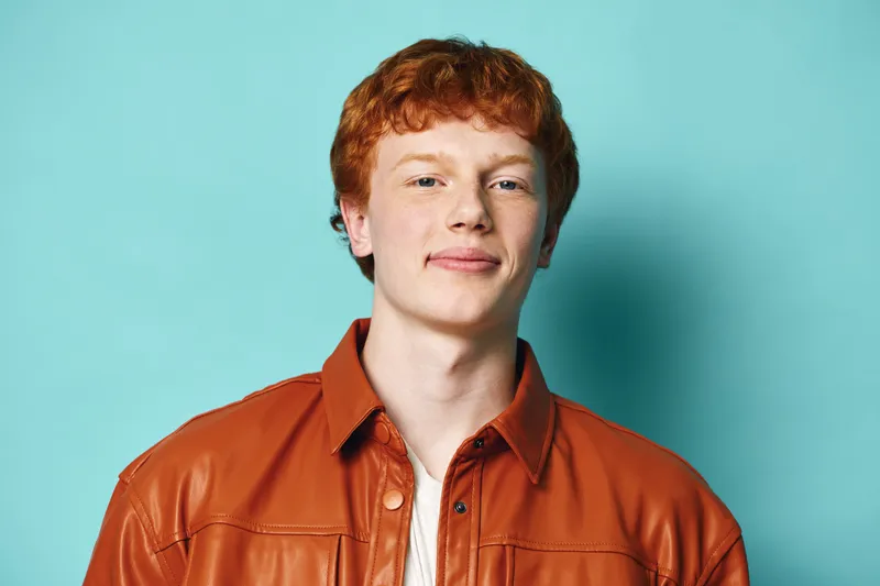 Young man with red hair and freckles wearing an orange leather jacket stands confidently against a blue background. Casual style and natural expression in portrait.
