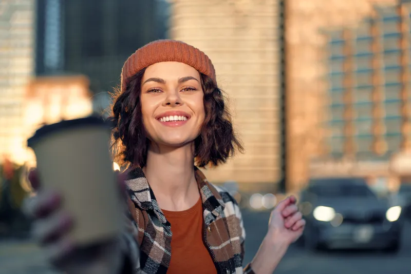 Woman smiling candidly holding coffee cup in urban street at golden hour, authenticity and lifestyle portrait with warm golden hour glow, mindful living and emotional storytelling vibe.
