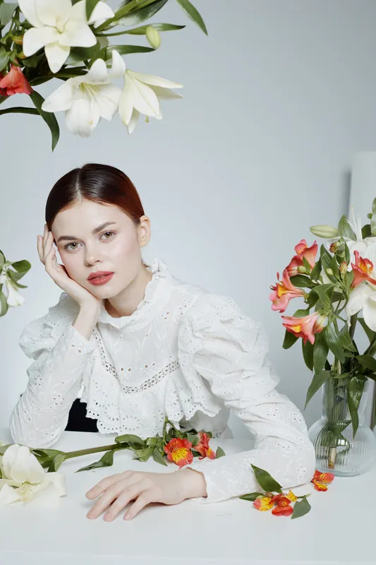 Elegant young woman in a white blouse surrounded by flowers, showcasing beauty and tranquility in a bright, minimalist setting