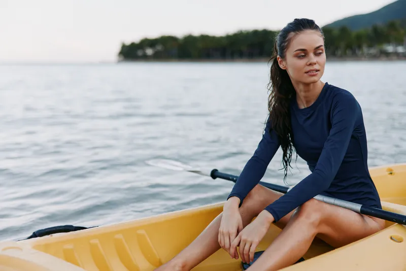 A young woman sits in a yellow canoe on calm water, wearing a dark blue long sleeve top, gazing into the distance as gentle light softens the scene.