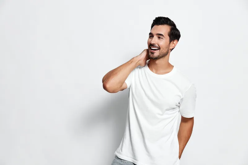 Smiling cheerful young man in casual white t-shirt poses playfully, looking at the camera with a friendly expression on a plain light background. People lifestyle concept.