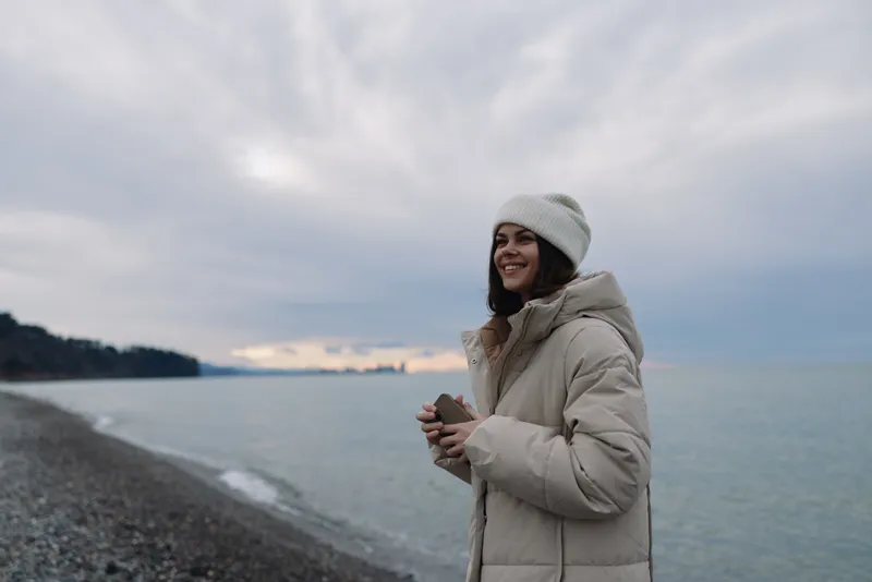Woman in a warm coat and knit hat on a windy beach, holding a phone, calm sea horizon under a cloudy sky, outdoor portrait on a chilly coastal day.