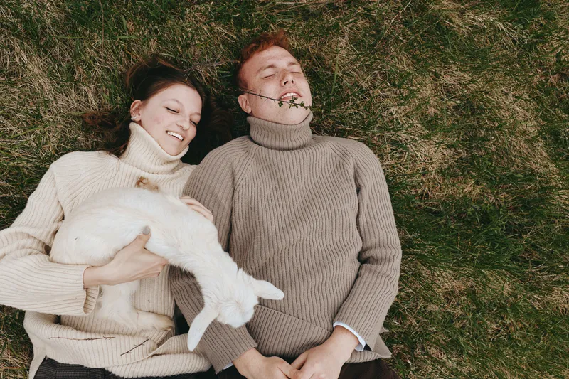 Candid lifestyle scene of a couple outdoors lying on grass with a playful goat kid nearby, cozy sweaters, sunlit moment, relaxed mood, natural colors, affectionate companionship
