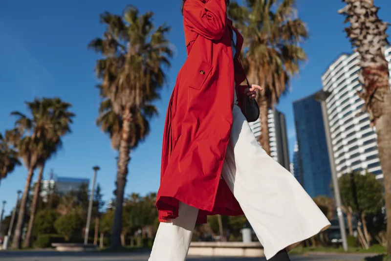 Young woman in a bright red coat walks briskly along a sunny city street, a white tote fluttering by her side as palm trees and modern high rises frame her lively motion.
