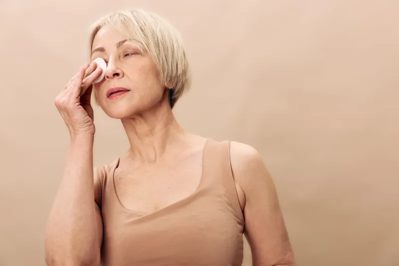 Mature woman with short light hair removes makeup with cotton pad on beige background. Concept of skincare, self-care, natural beauty, and daily facial cleansing routine for aging skin.