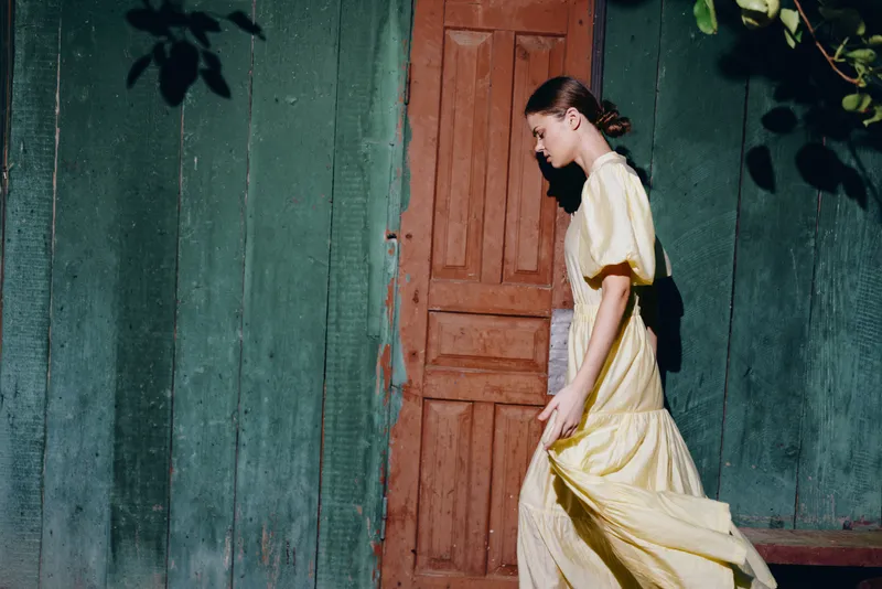 Woman wearing a yellow dress walks past a weathered doorway along a green wall exterior, sunlight casting soft shadows and creating a timeless vintage outdoor portrait.