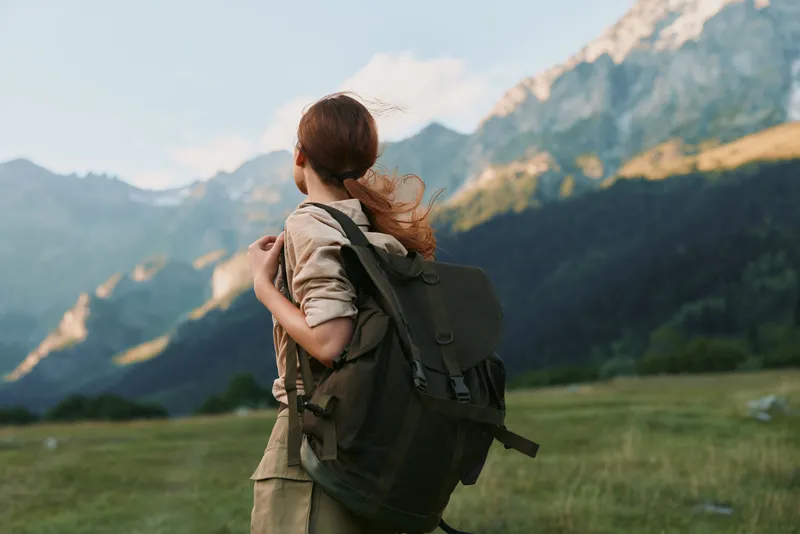 Woman lifestyle traveler with backpack standing on green field looking at mountain landscape under blue sky, outdoor adventure, nature exploration, casual clothing