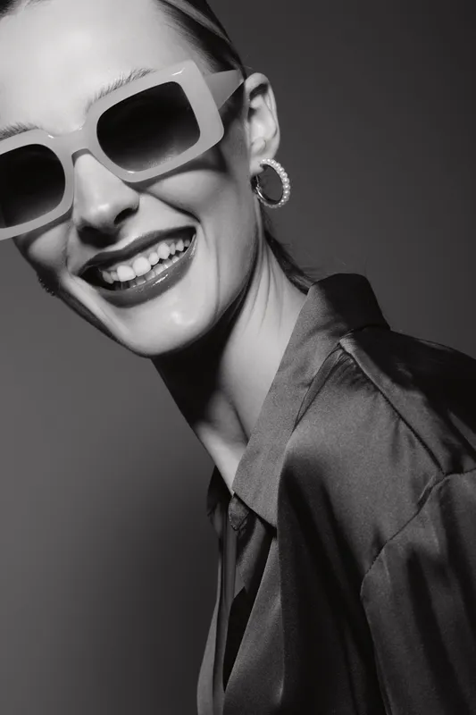 Smiling caucasian woman with light skin wearing large sunglasses and hoop earrings in satin blouse. Studio portrait in black and white with soft lighting and joyful mood.