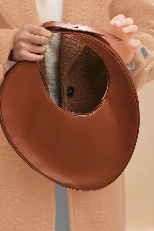 Asian woman holds a round brown leather handbag in a chic coat, presenting fashion detail with poised confidence. The image conveys a stylish, modern lifestyle and subtle elegance.