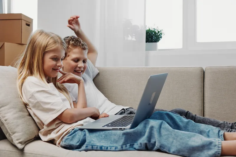 Two children lying on sofa using laptop together in cozy living room. Happy kids smiling and enjoying digital entertainment and learning at home.