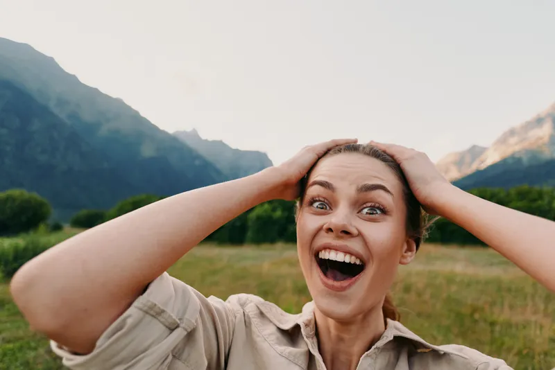 Young woman outdoors in a sunlit field with mountains in the background, holding her head with a wide smile, casual beige shirt, wind in her hair, natural colors.