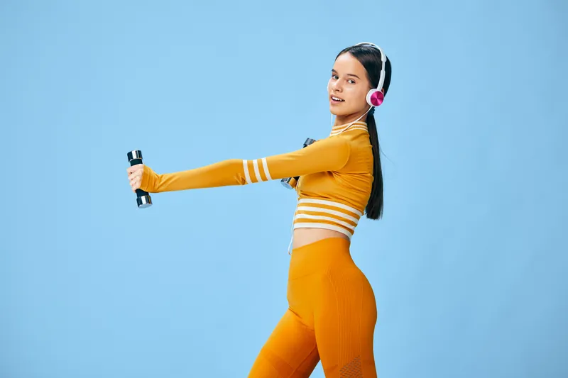 Fitness oriented portrait of a woman in bright orange activewear, extending arms with dumbbells, wearing pink headphones against a blue studio backdrop for energetic sport and workout visuals.