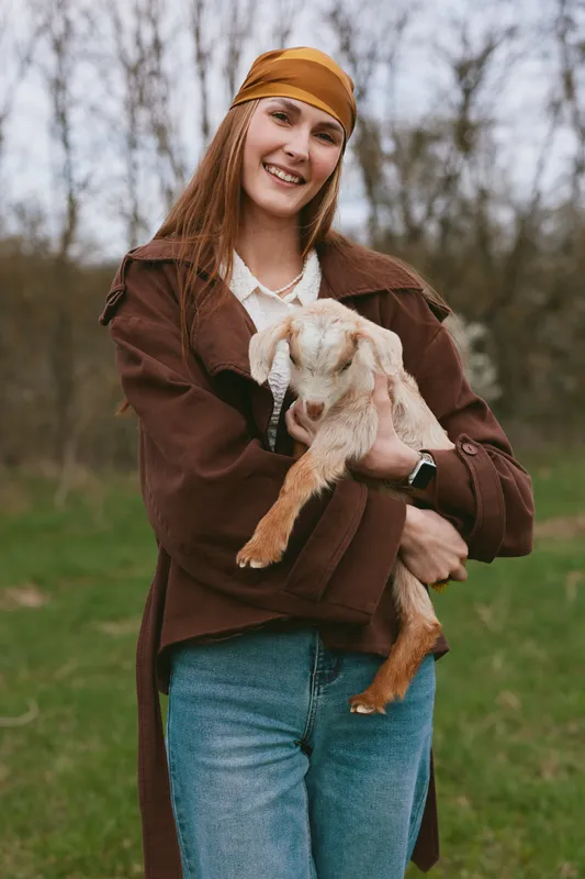 Joyful woman outdoors with a goat kid, sharing a warm moment in a rustic lifestyle setting. A candid scene of care, companionship, and natural farming vibes in a grassy field.