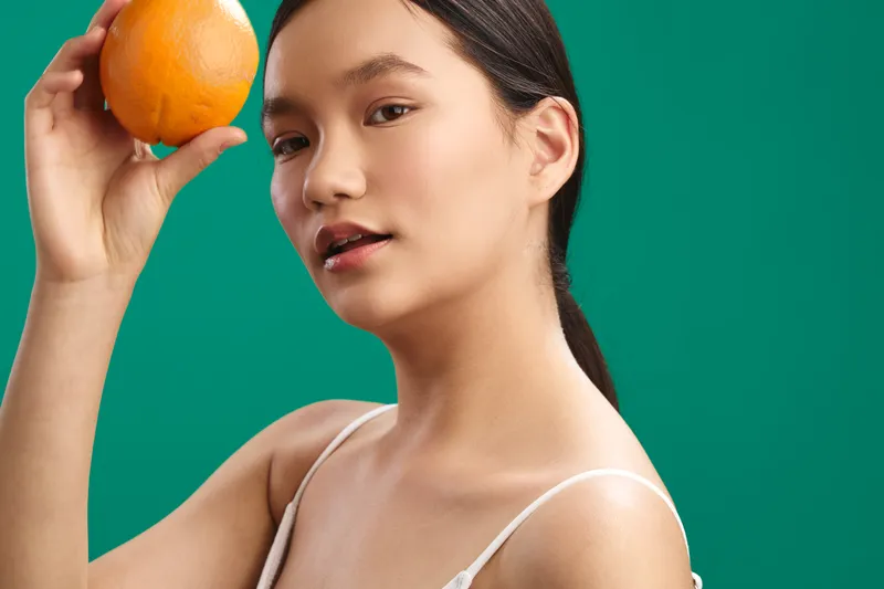 asian woman holds an orange near her head in a clean studio with a vivid green background, a calm confident expression, and a fresh energetic mood that feels natural and inviting.