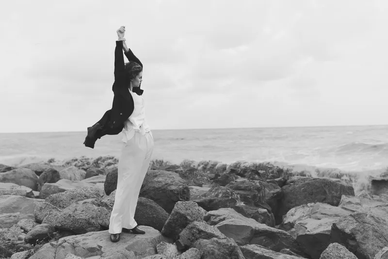 Woman stands on rugged rocks by the sea, arms raised in triumph, wind catching her jacket and white pants, a monochrome outdoor moment that highlights freedom, resilience and coastal adventure along
