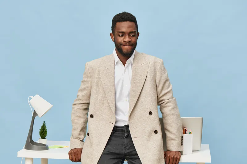 Stylish happy man wearing beige blazer and white shirt standing confidently in modern office with desk and laptop, blue background, business casual look, modern workplace setup, professional lifestyle
