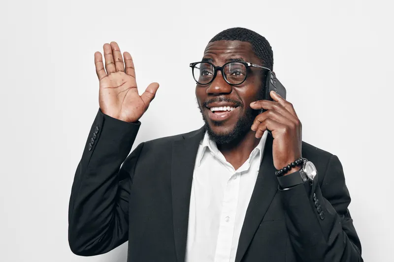 African American man with dark skin wearing glasses and suit talking on phone and waving hand with smile on white background for communication and greeting.