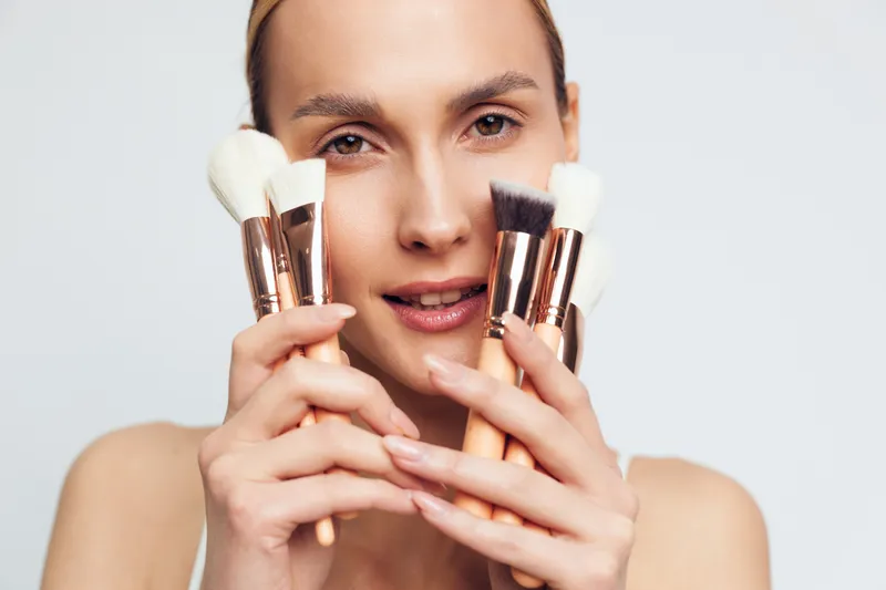 A young adult female holds makeup brushes up to her face in a clean studio, highlighting cosmetics application and a focused, preparatory mood before a beauty session.