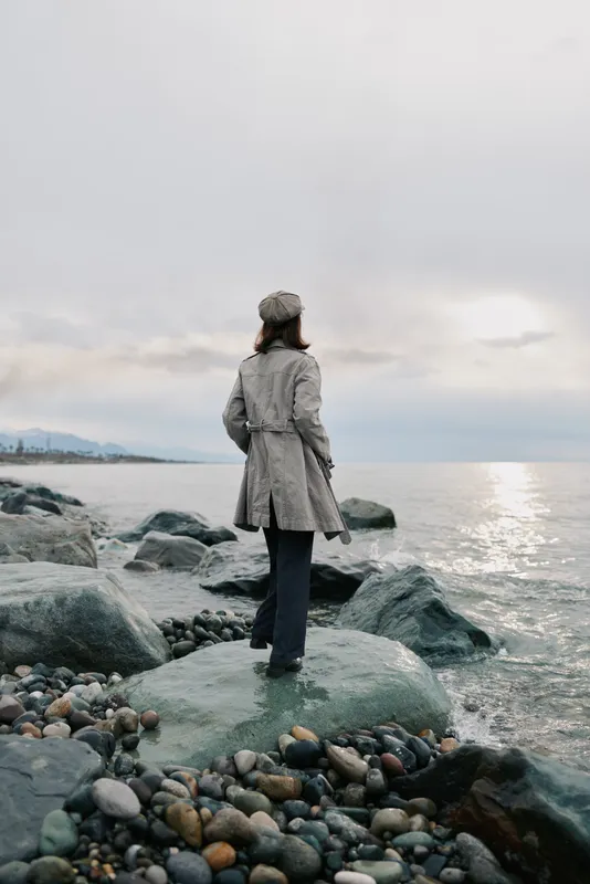 A woman in a gray coat and hat stands on a rock by the sea, looking at the calm water under a cloudy sky. The rocky shore creates a peaceful, reflective mood.