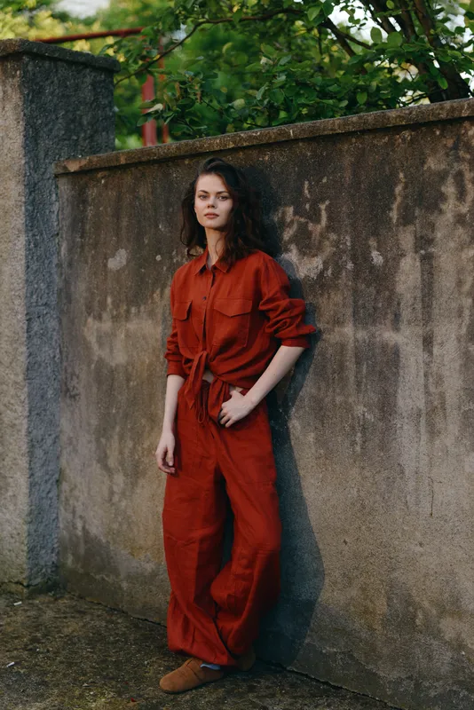 Confident woman outdoors in a red jumpsuit leaning against a mossy wall, natural light highlights her relaxed stance, urban vibe, candid lifestyle moment with breeze