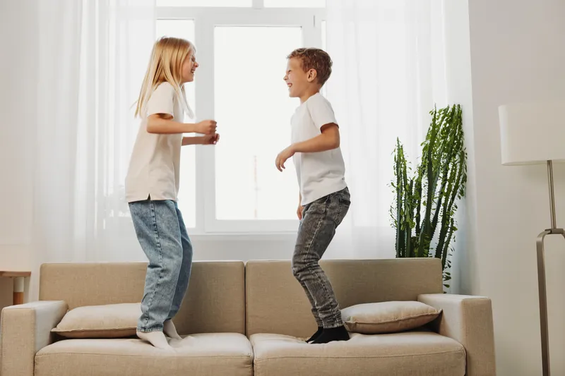 Two happy children jumping on a beige sofa in a bright living room with natural light. The casual outfits and joyful expressions show playful energy and carefree childhood moments.