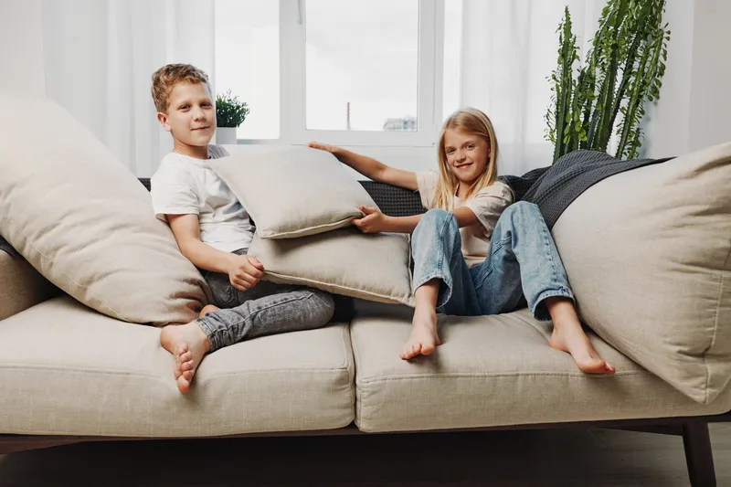Two happy children play pillow fight on beige sofa in bright living room with plants. Casual home atmosphere with laughter and sibling fun.