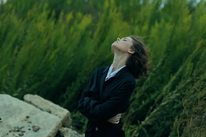 Businesswoman in a dark suit stands among rocks and greenery, head tilted back, arms crossed, embracing breeze and reflection in a natural setting.