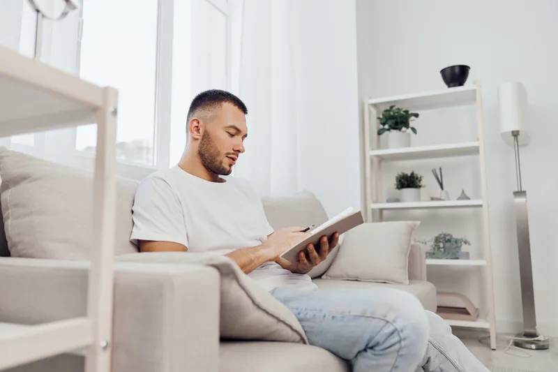 Thoughtful man reading a book on a cozy couch, surrounded by modern decor and soft natural light, reflecting comfort and relaxation in a contemporary living space.