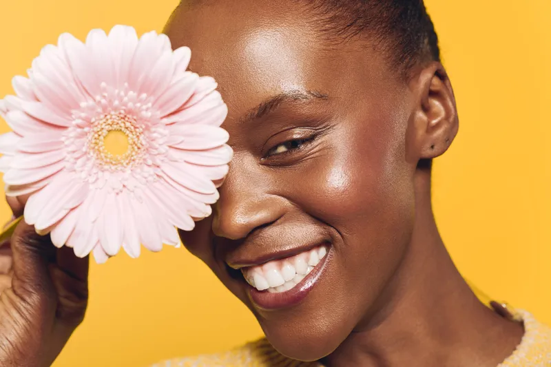 Smiling black woman with dark skin holds pink flower near face on yellow background. Concept of beauty, skincare, happiness, natural glow, and floral wellness in studio lighting.