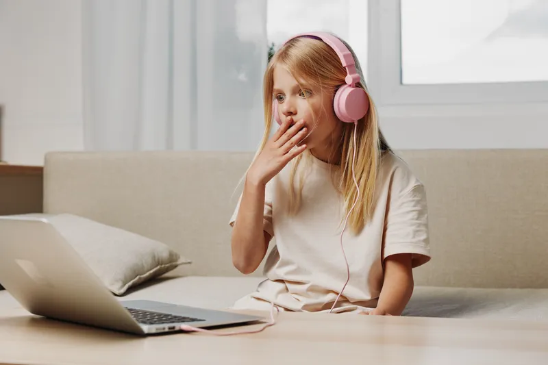 Curious girl with pink headphones watches laptop screen surprised, sitting on couch in bright modern room. Concept of online learning and childhood curiosity.