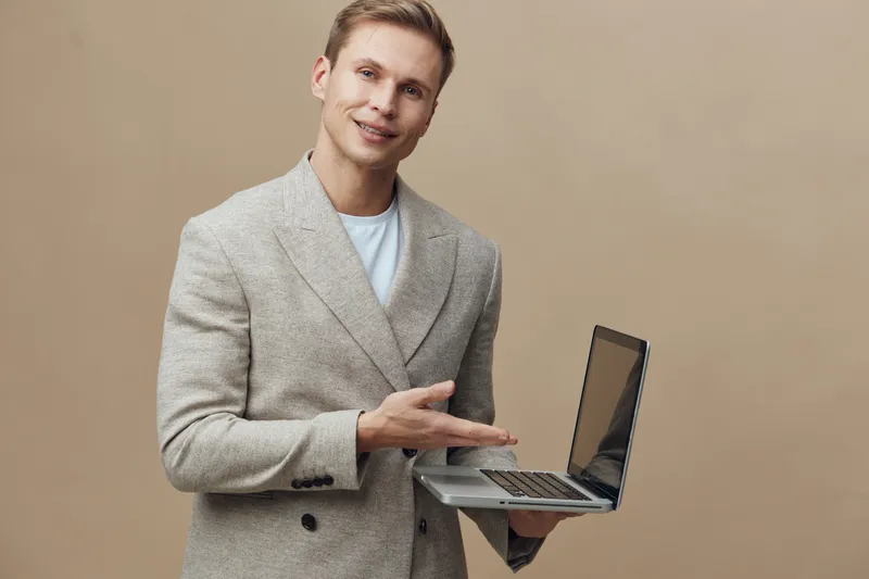 Smiling man holding laptop computer in hand looking at camera, casual business attire, indoor studio background, light beige color, professional technology workplace concept.