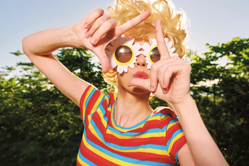 Playful woman outdoors wears colorful striped shirt and round sunflower sunglasses, capturing a retro film color vibe with vibrant mood, a candid portrait against bright sky and green foliage.