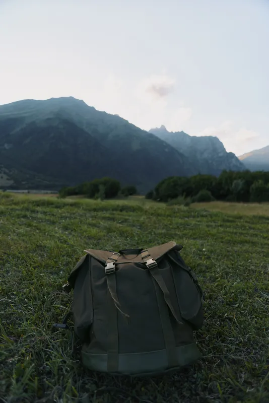 Green backpack stands alone on grassy field with mountain range in the background under a cloudy sky during daytime.