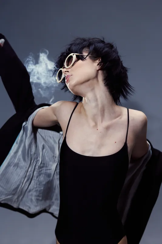 Stylish woman in black clothing with sunglasses blowing smoke, edgy and modern vibe with dark background.