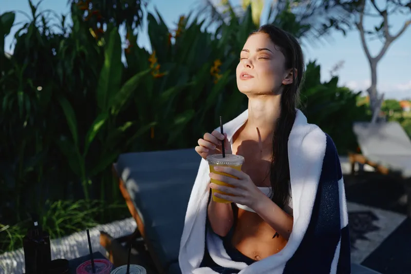 A sunlit poolside scene shows a woman wrapped in a towel, savoring a cold beverage with closed eyes. A moment of relaxation, summer calm, and outdoor luxury by the bright pool.