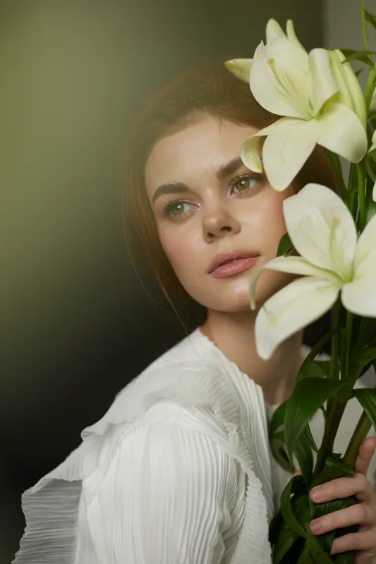 Beautiful young woman with lilies in a soft light, wearing a white blouse, exuding calmness and elegance, perfect for floral or beauty concepts