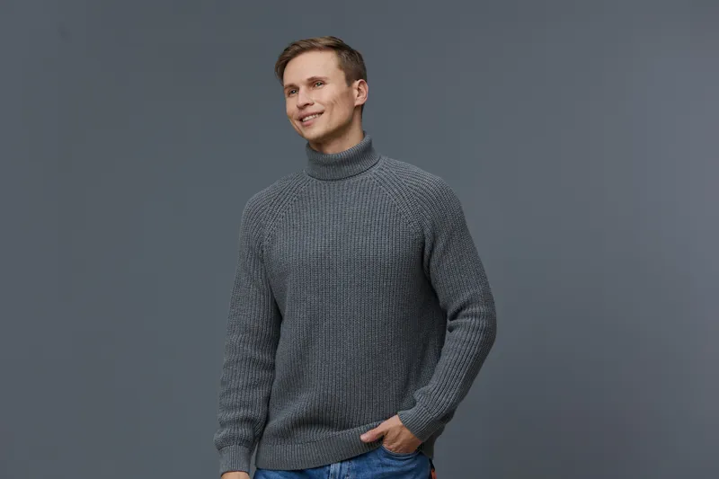 Smiling man wearing gray knitted turtleneck sweater on plain gray background, casual fashion, cheerful mood, studio portrait, fashion clothing, lifestyle concept, modern style.