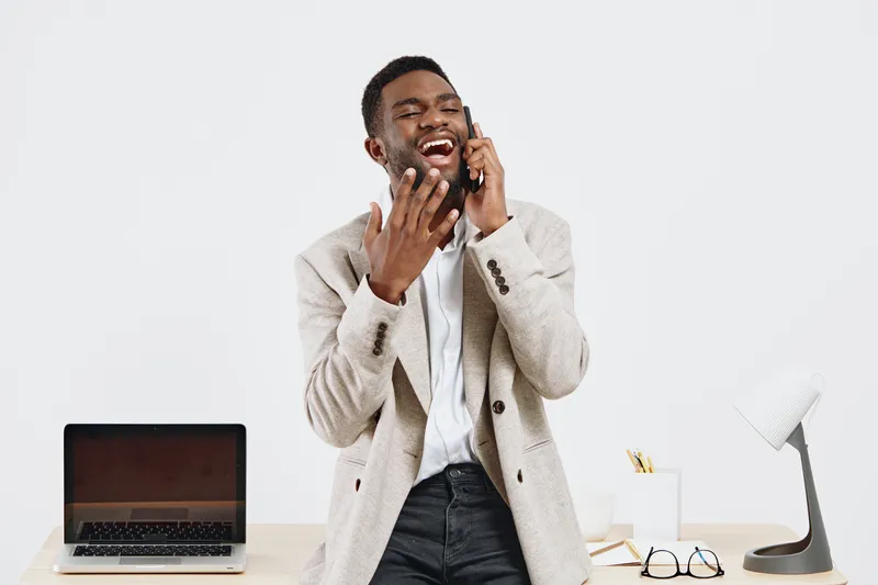 Bright cheerful young African American man with beard wearing beige blazer talking on mobile phone while smiling and looking happy, indoor modern office setting, lifestyle concept.