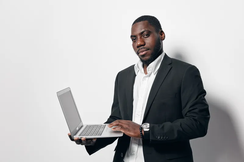 African man with dark skin holding laptop and wearing black suit and white shirt standing against white background for business or technology concept.