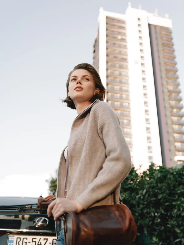 A poised young woman stands beside a car on a city street, gazing upward with a calm, determined expression, wearing a neutral coat and carrying a leather bag.
