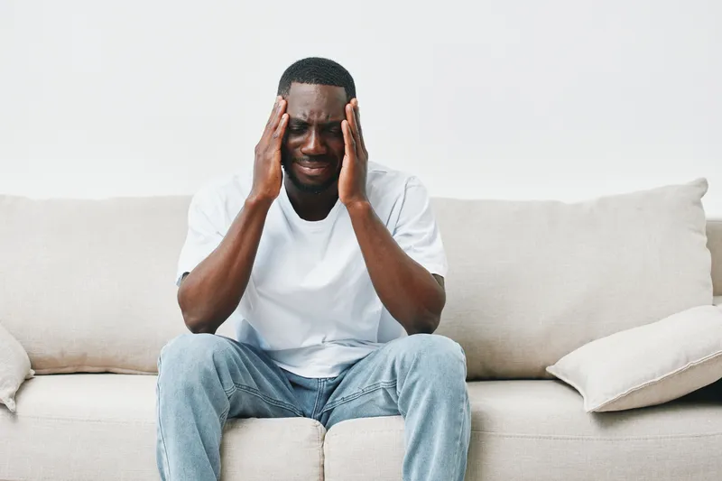 Emotional portrait of a young Black man sitting on a couch, experiencing stress and anxiety, wearing a white t shirt and jeans in a minimalist living room setting